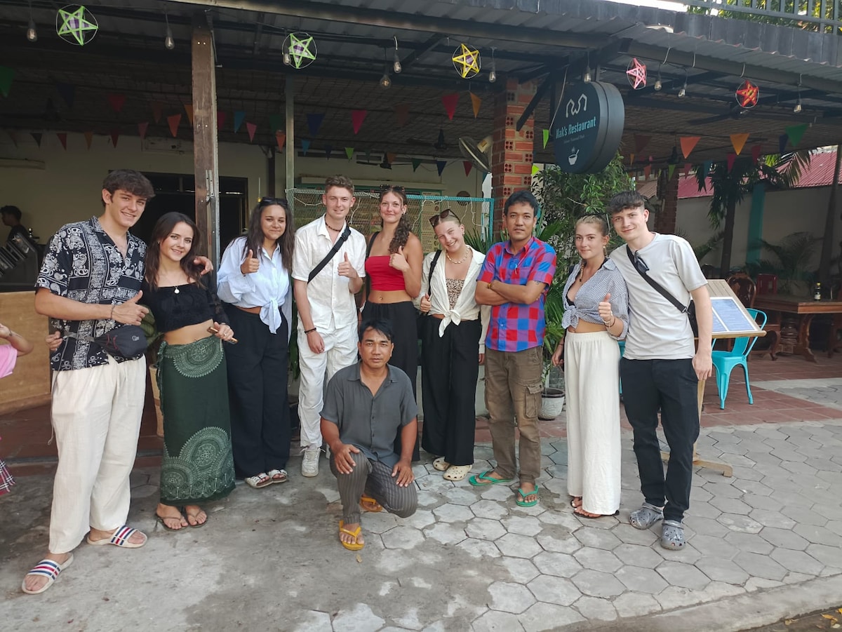 A diverse group of individuals stands in front of a guesthouse, dressed in various styles of clothing. The setting features a covered outdoor area adorned with colorful decorations. The pathway is made of patterned tiles, and several chairs are visible in the background.