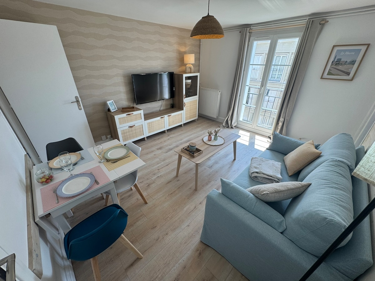 A well-lit living area features a comfortable blue sofa facing a modern media unit with a television. A dining table for two is set with dishes and a small bowl of fruit. Large windows allow natural light to fill the space, accentuated by light-colored curtains.