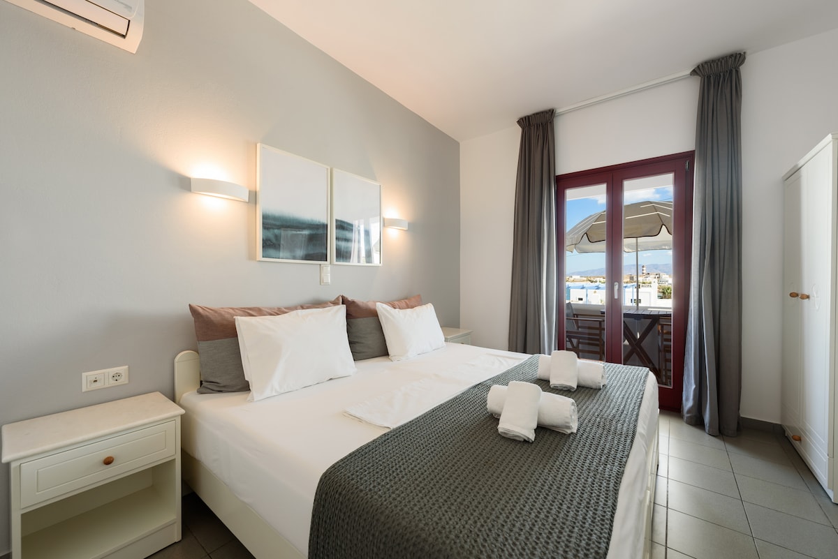 A spacious sleeping area features a light-colored bed adorned with neutral-toned pillows and a textured throw. Two framed art pieces are mounted on the wall above the bed. Natural light enters through a set of double doors, which lead to a small balcony.