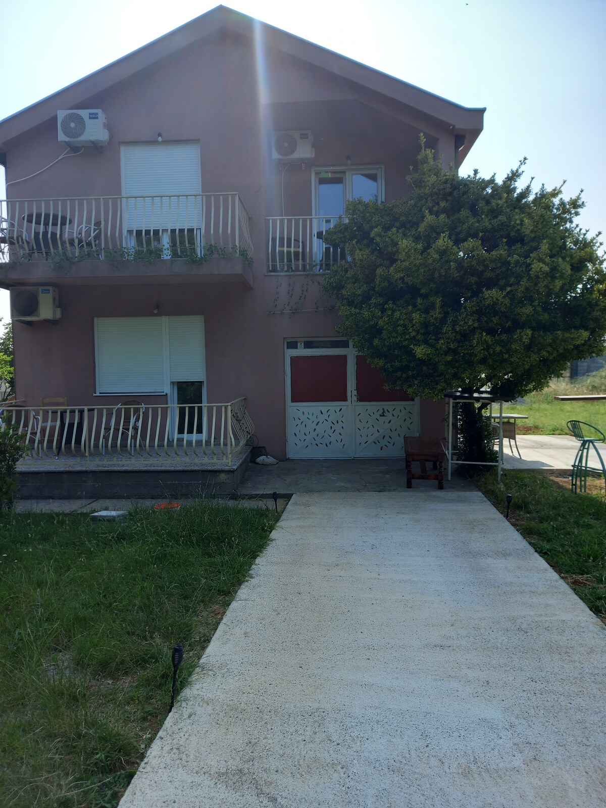 The exterior of a two-story building is presented, featuring a simple design with balconies and two air conditioning units. A light-colored pathway leads to the entrance. Lush greenery surrounds the property, including a bushy tree visible nearby.