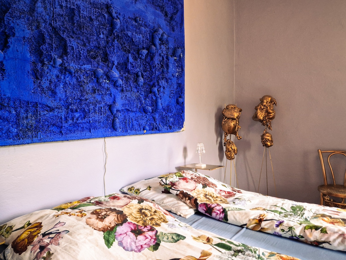 A serene sleeping area is highlighted by a vibrant blue wall art piece. Floral bedding adorns the spacious beds, while unique sculptural art stands nearby. A minimalist bedside table with a glass sits next to the beds, creating a harmonious and artistic setting.