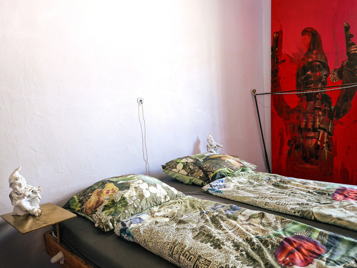 A bedroom displays two beds adorned with floral-patterned bedding. A small metallic table with a sculptural figure is positioned beside one bed. A striking red wall features a large artwork, contributing to the eclectic atmosphere of the room.