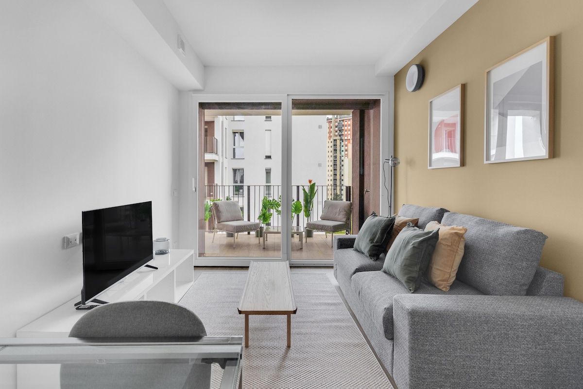 A modern living room is displayed, featuring a spacious gray sofa with decorative pillows and a low wooden coffee table. Large sliding glass doors lead to a balcony, where outdoor seating is surrounded by green plants, allowing natural light to brighten the space.