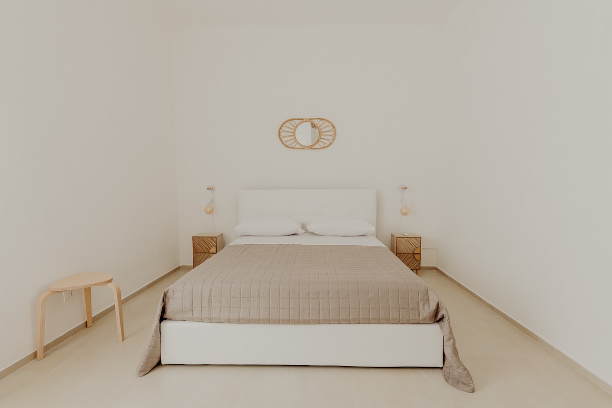 A spacious bedroom features a large bed with neutral bedding. Nightstands are placed on either side, each holding a lamp. A decorative mirror adds interest to the light-colored wall, while the overall minimalist aesthetic enhances a calming atmosphere.