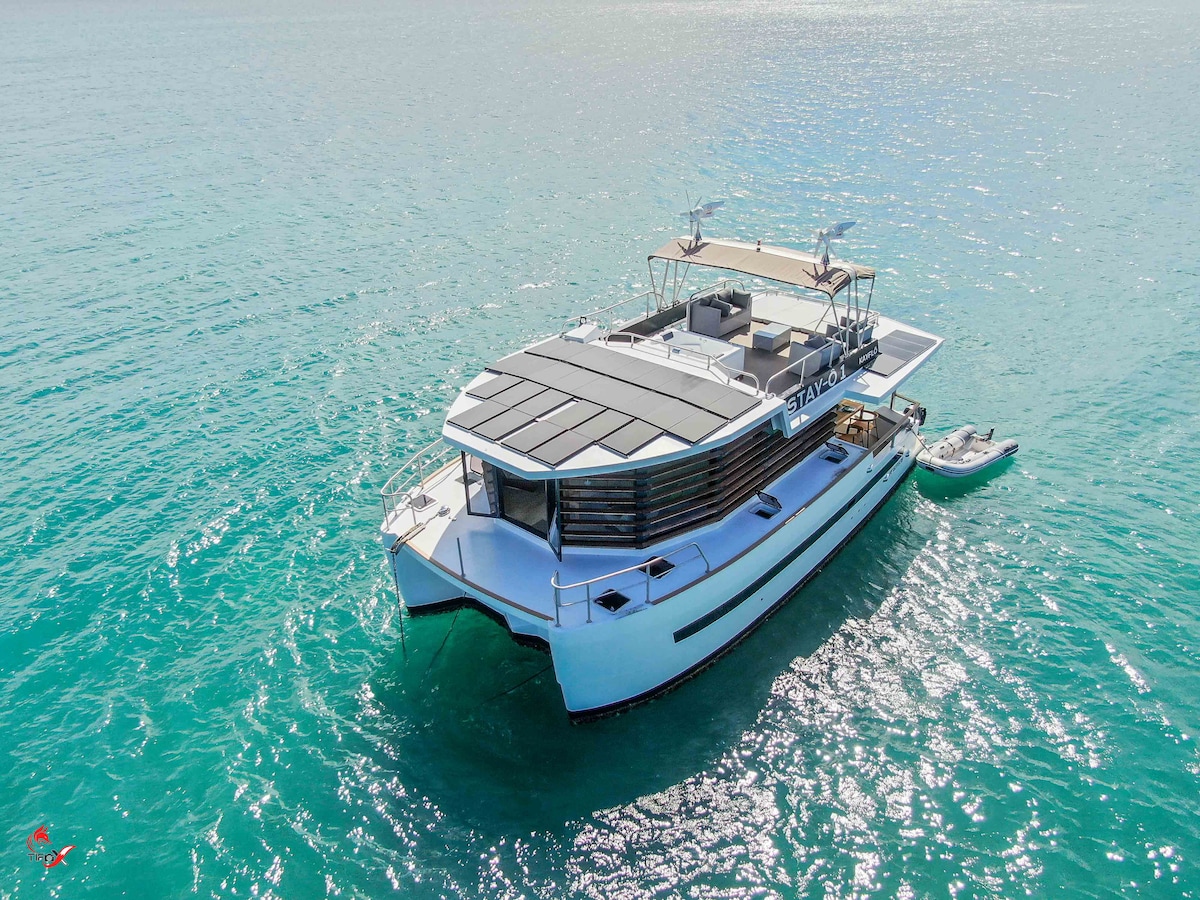 The eco-friendly floating lodge is showcased from above, surrounded by clear turquoise waters. Its modern design features clean lines and large windows. A spacious deck is visible on the upper level, accommodating outdoor seating and sunbathing areas, with a small boat anchored nearby.