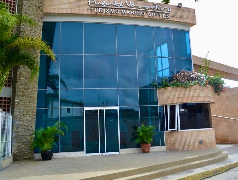 The entrance of a modern building features expansive glass windows that reflect the surrounding greenery. A curved awning extends over the glass doors, while potted plants add a touch of nature to the entrance area.