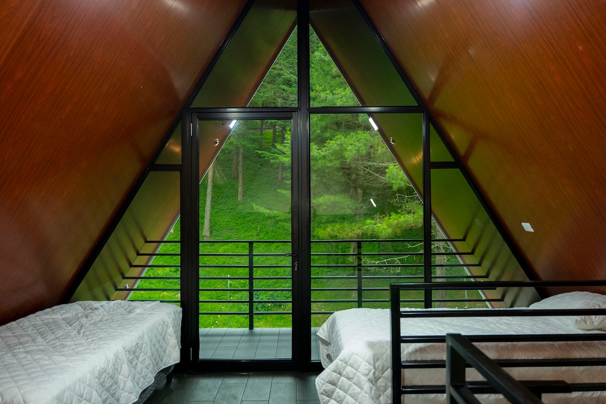 A cozy sleeping area is situated in the upper part of the cabin, featuring two twin-size beds with light-colored bedding. Large windows offer a panoramic view of the surrounding greenery, while the triangular wooden walls enhance the cabin's rustic charm.