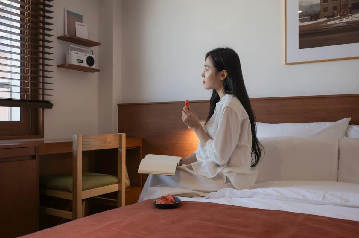 A serene hotel room features a spacious bed with crisp white linens and a warm wooden headboard. A person is seated at a writing desk, enjoying a snack, while natural light filters through the window's wooden blinds. Soft tones create a calm atmosphere.
