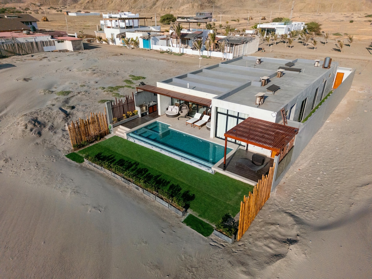 An aerial view of a modern luxury house reveals a private pool surrounded by a well-maintained lawn. The outdoor area features lounge chairs and shaded seating, set within a tranquil desert landscape. The structure displays a contemporary design with large windows and an inviting entrance.