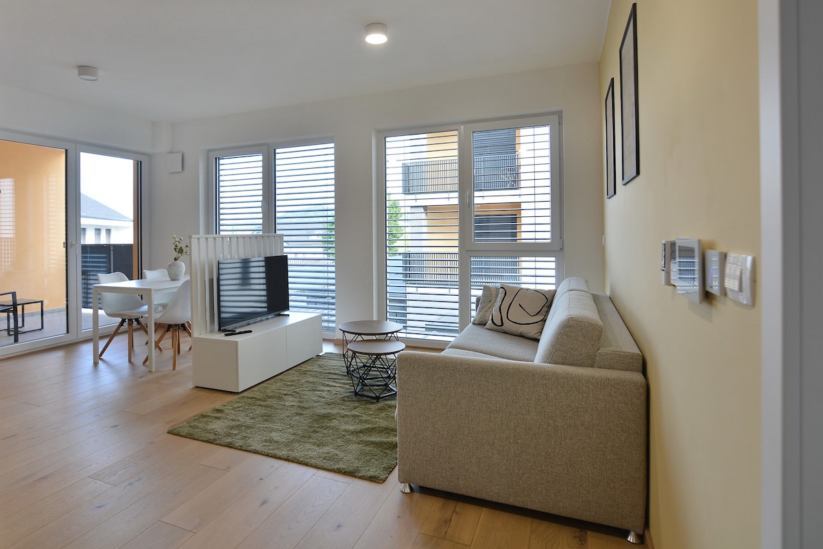 A living area features a light-colored couch positioned next to a small coffee table on an area rug. A flat-screen TV is mounted on a white media console, and large windows allow natural light to fill the space, while offering views of the outside terrace.