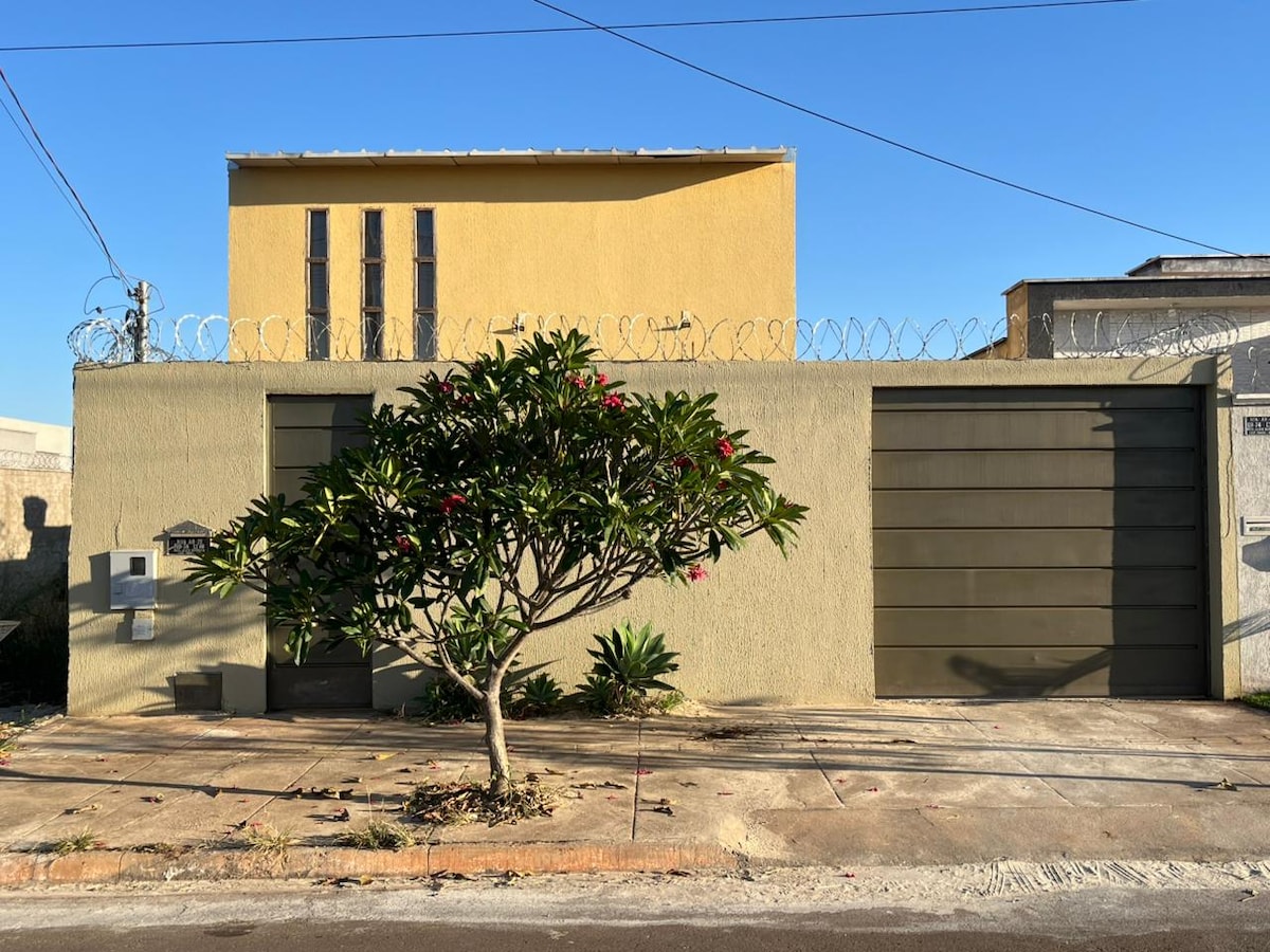 The exterior of the house features a yellow façade with a modern architectural design. A small tree with pink flowers is positioned in front of the property. Barbed wire lines the top of the wall, and a large garage door is visible on one side.