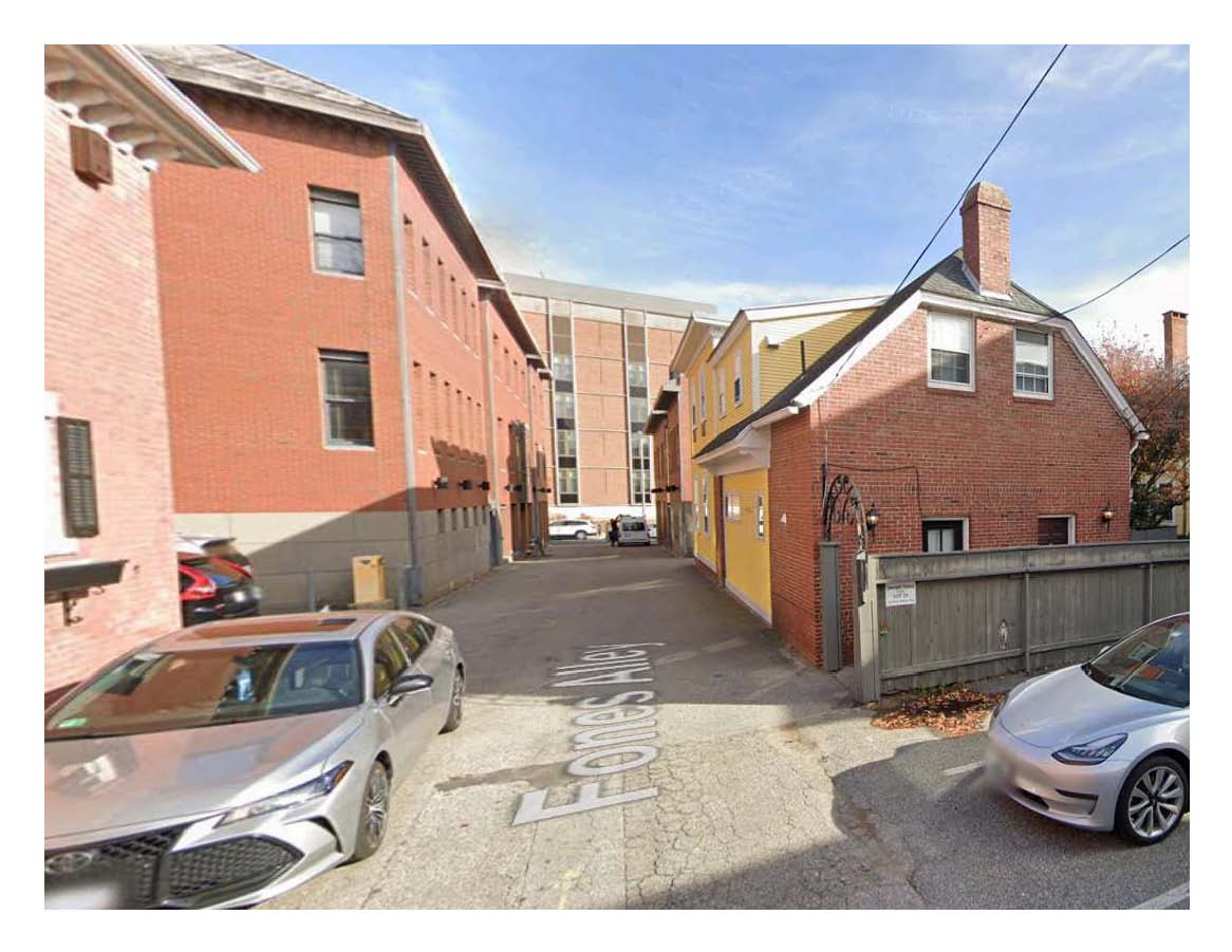 A narrow alleyway is framed by red-brick buildings on the left and a yellow building on the right. Parked cars are visible along the left side, with a wooden fence suggesting an enclosed area at the end of the alley. The sky is clear and bright.