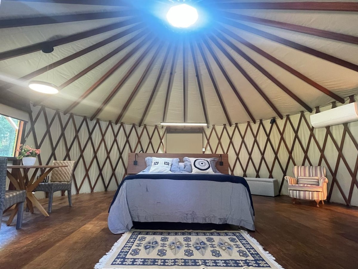 An interior view of the yurt shows a spacious sleeping area with a large bed centered beneath a circular skylight. Light-colored walls are complemented by wooden beams, while a patterned rug sits beneath the bed. A striped armchair and a small table with woven chairs are also present.
