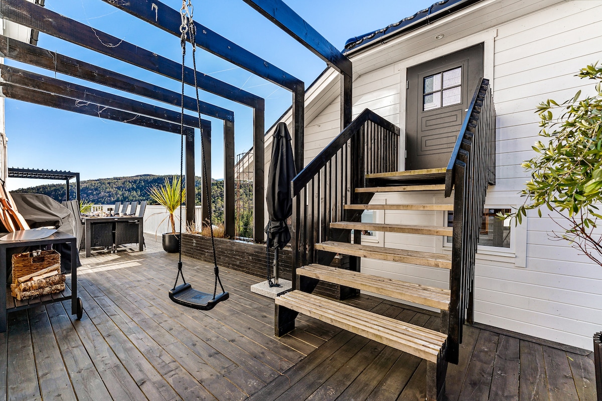 An outdoor deck features a wooden staircase leading to the entrance. A swing is suspended from the overhead beams, while seating areas are visible in the background. Potted plants and a grill contribute to the inviting ambiance of the space.