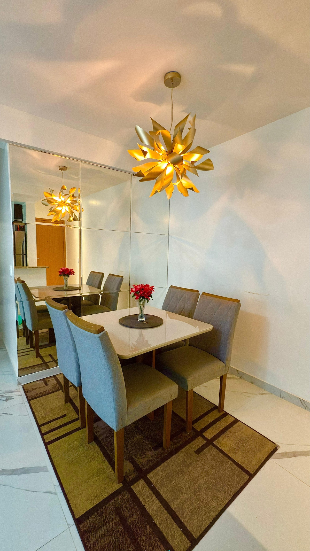 A dining area is presented with a white table surrounded by six gray upholstered chairs. A decorative center piece of red flowers is placed on the table. A striking light fixture with petal-like design hangs above. Mirrored walls reflect the space, enhancing its brightness.