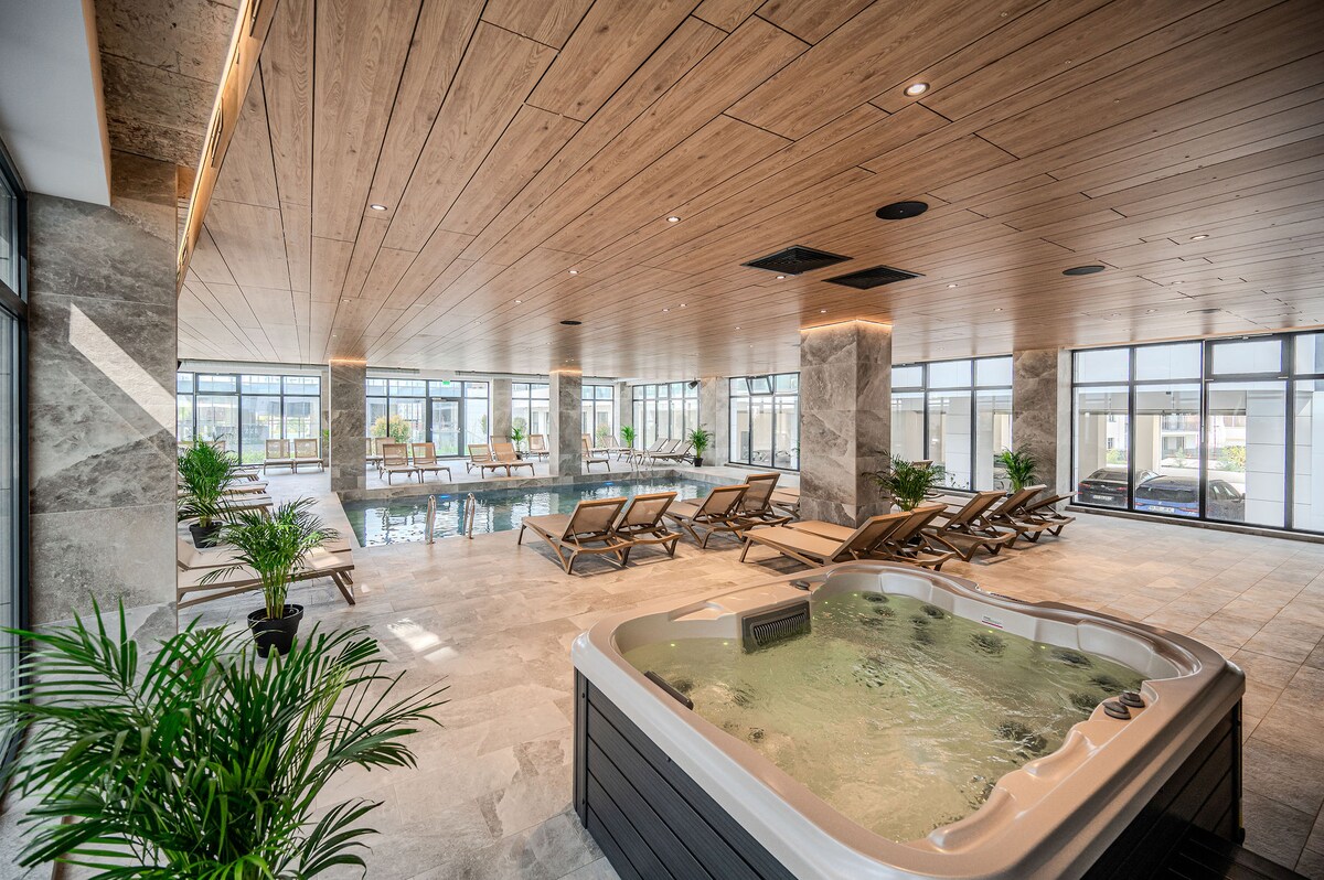 An indoor wellness area features a spacious swimming pool surrounded by comfortable lounge chairs. A hot tub with bubbling water is situated nearby, complemented by greenery in planters. Large windows allow ample natural light to fill the area.