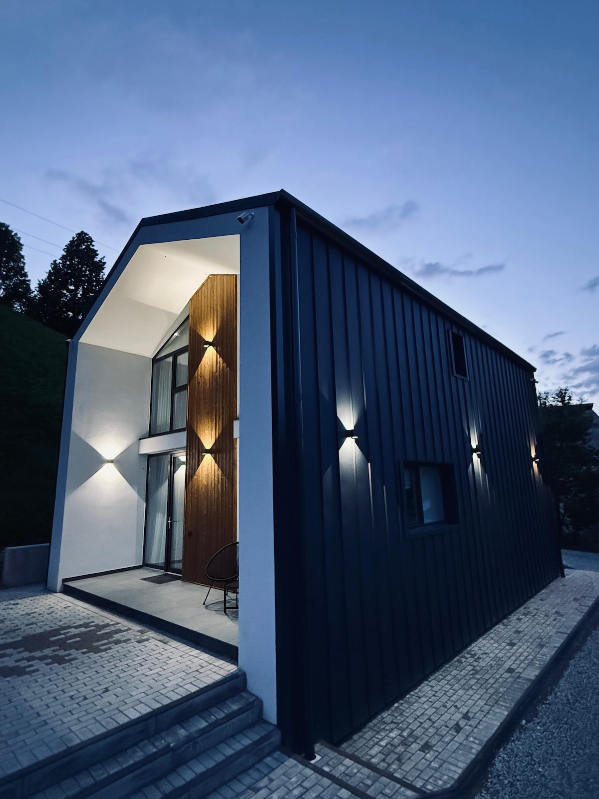 The modern exterior of the house is illuminated at dusk, showcasing a striking geometric shape. Smooth, dark siding contrasts with warm wooden accents around the entrance, framed by subtle lighting that highlights the architectural details.
