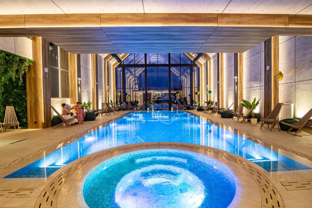 An indoor pool area is illuminated by soft lighting, with a long lap pool at the center. A hot tub is positioned nearby, surrounded by modern lounge chairs. Large windows offer views of the outside, while tropical plants add a touch of greenery to the space.