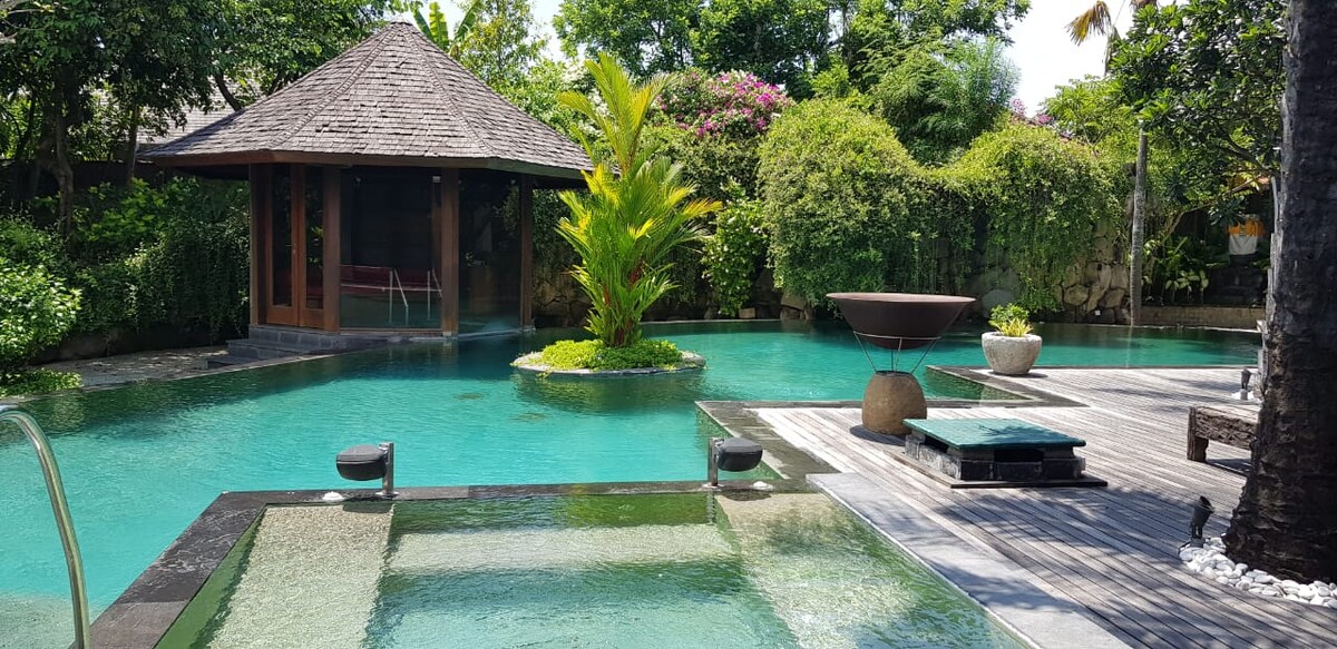 A serene pool area features a large, inviting swimming pool surrounded by lush greenery. A wooden pavilion with a thatched roof is visible in the background, while smooth stones and well-manicured plants enhance the landscape. Reflections on the water create a calming atmosphere.