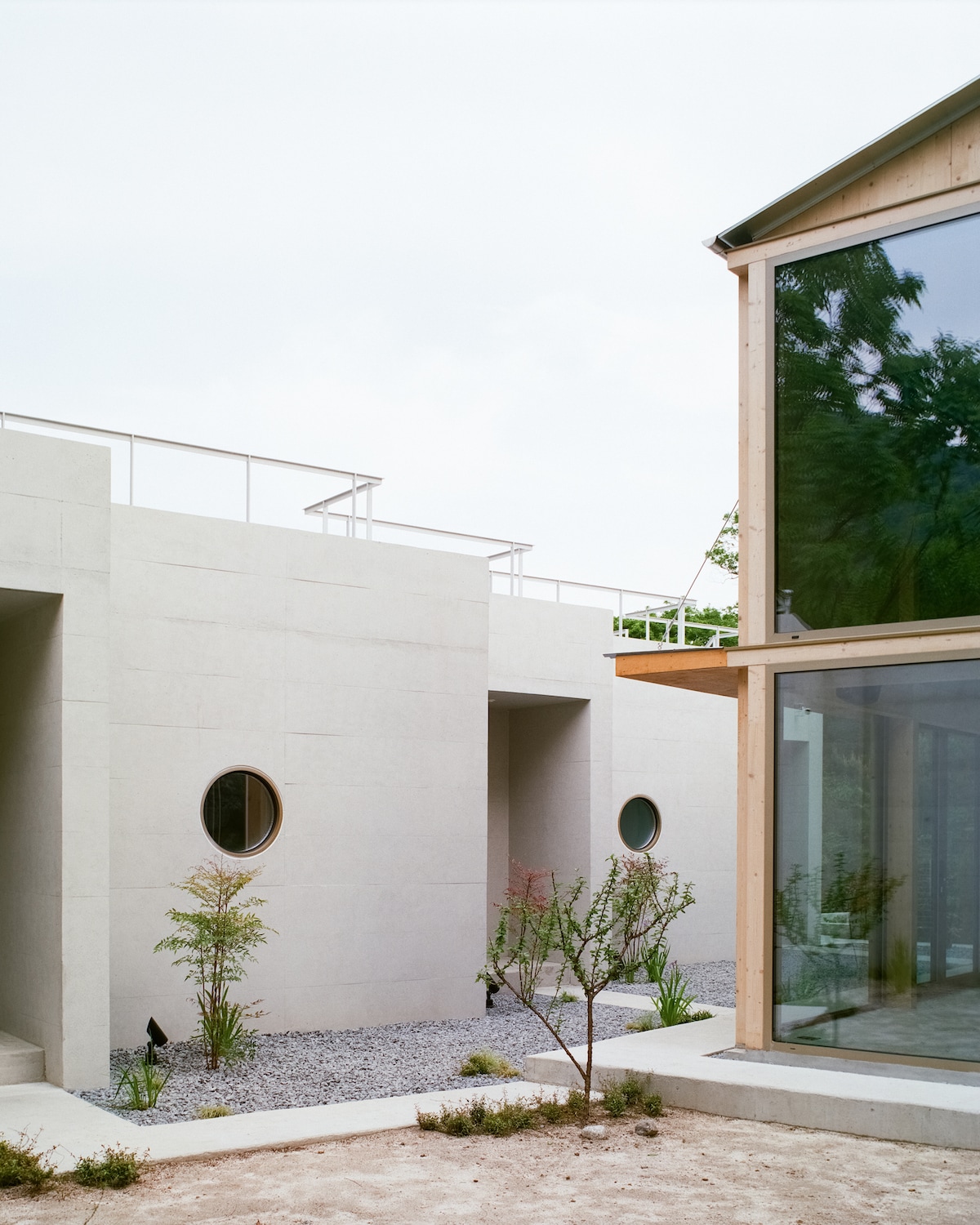 The image features a modern architectural exterior with smooth, light-colored walls and large windows. A rooftop area is visible, alongside circular windows on the structure. Sparse landscaping with small plants and gravel pathways creates a serene environment, inviting contemplation and relaxation.