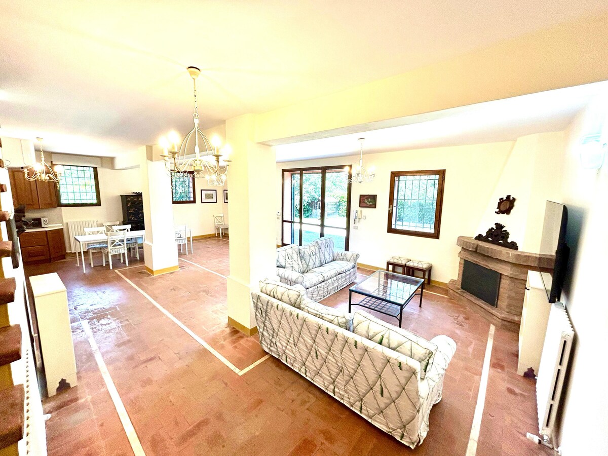 A spacious living area features two comfortable sofas positioned around a coffee table. Natural light fills the room through large windows, offering views of the surrounding greenery. An open dining space with a white table and chairs is visible in the background, along with a cozy fireplace.