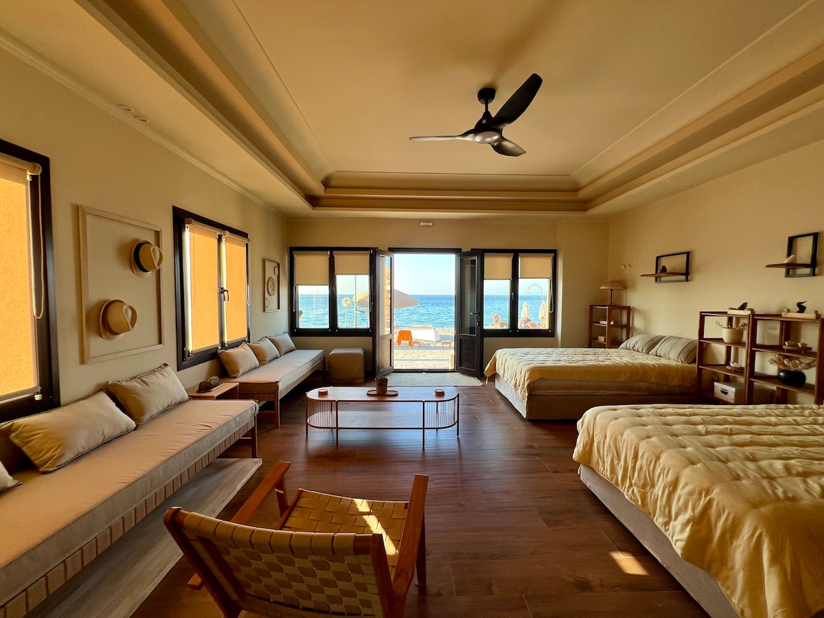 A spacious room features large windows allowing natural light and a view of the nearby beach. Two comfortable beds are arranged alongside a seating area with a sofa and a chair. Decorative elements, including hats and wooden shelves, add charm to the welcoming atmosphere.