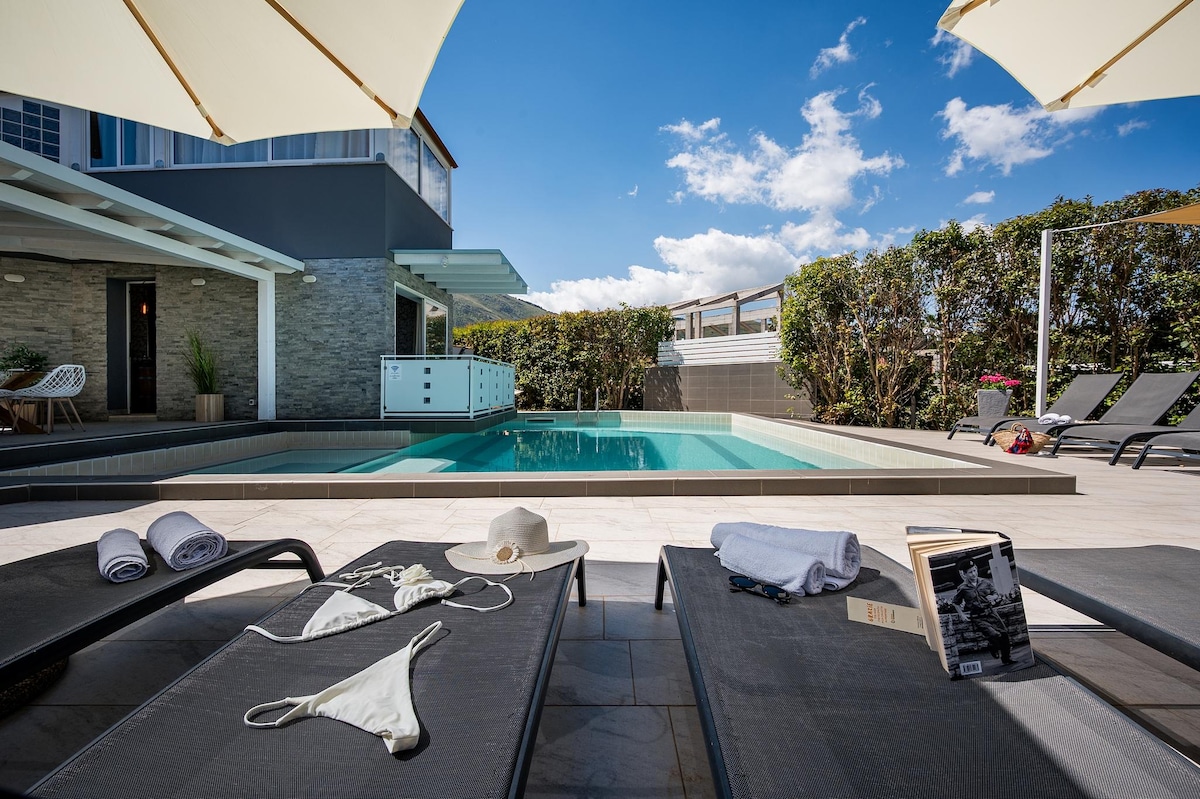 Lounge chairs are positioned around a private pool, with sun umbrellas providing shade. The pool is surrounded by a tiled deck and lush greenery. A few personal items, including towels and a sun hat, are placed on the chairs, suggesting a relaxed outdoor space.