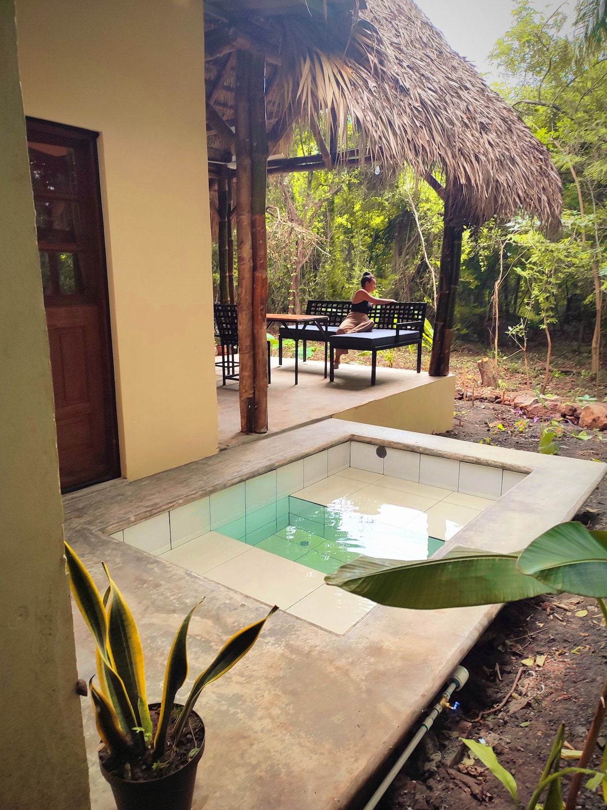A private terrace is shown with a thatched roof overhang, providing shade to seating. A small plunge tub is visible, partially filled with water, surrounded by natural greenery. The tranquil atmosphere is enhanced by the adjacent plants and the seating area's view of the jungle.