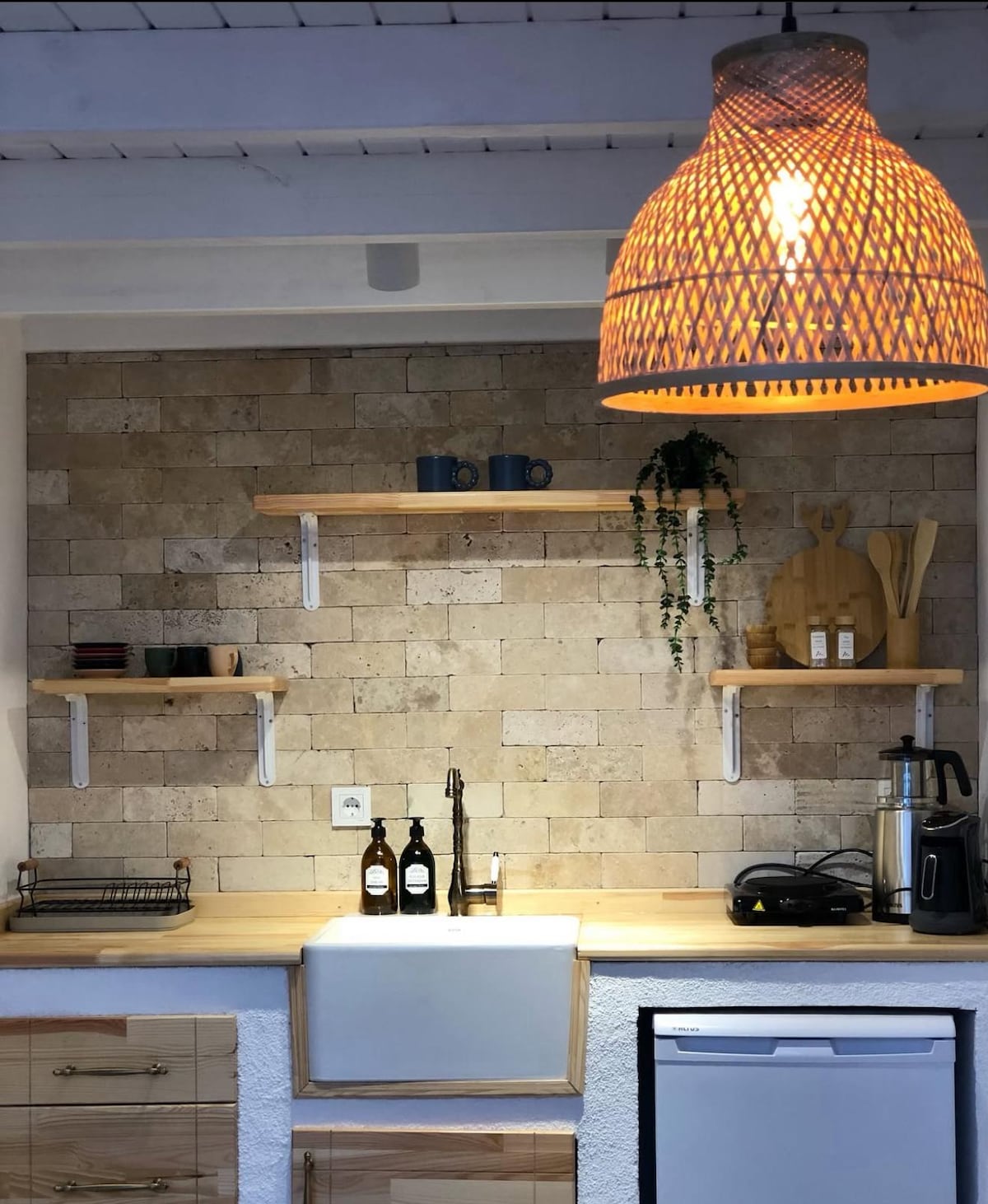 A modern kitchen space is highlighted by a textured stone wall and light wooden shelves. A large sink is positioned below the shelves, accompanied by decor items, utensils, and cooking essentials. A wicker pendant light hangs above, providing soft illumination.