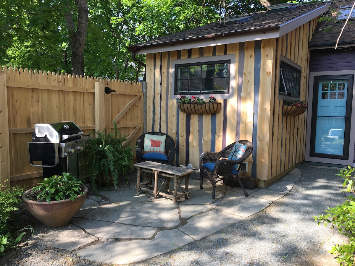 A private patio area features a stone pathway surrounded by greenery and planters. A small wooden table is flanked by two chairs, offering a cozy seating arrangement. A barbecue grill is positioned against a wooden fence, providing an outdoor cooking option.