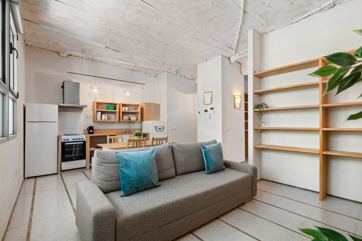 A modern living space features a light grey sofa adorned with blue cushions, positioned in a spacious area. Open shelves line the wall, providing ample storage. A functional kitchen is visible, equipped with essential appliances, all highlighted by natural light from large windows.