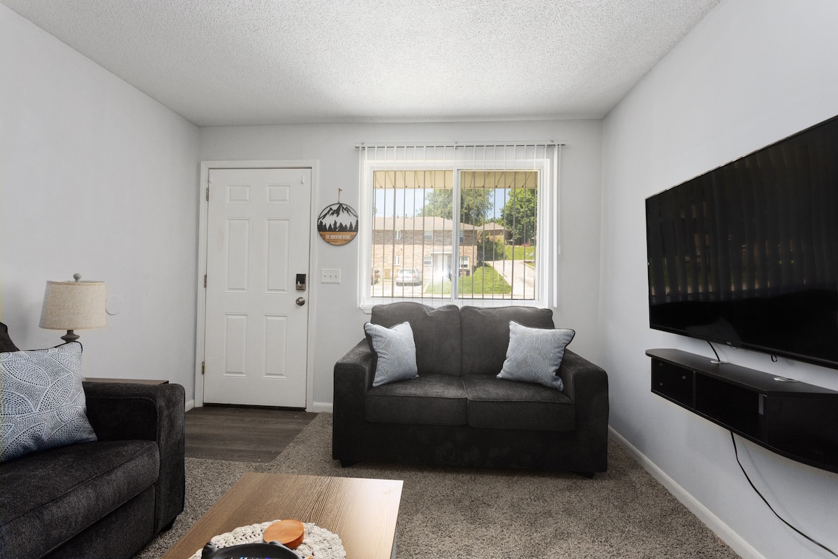 A simple living area features a dark sectional sofa and a light coffee table. Natural light enters through large windows, illuminating the room, while a flat-screen television is mounted on the wall opposite. A decorative charm hangs beside the front door.
