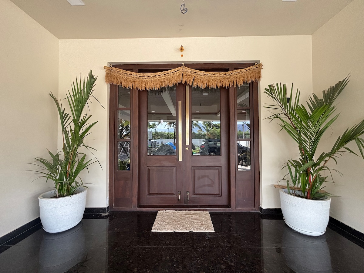 Double wooden doors, framed with decorative tassels, welcome guests into the home. On either side, large white pots hold vibrant green plants. A neutral-colored mat lies on the polished dark floor, enhancing the serene entryway ambiance.
