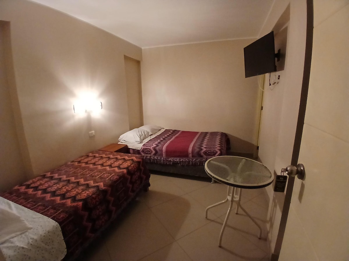 The room contains two beds, one with a patterned cover and the other with a crisp white sheet. A small round table is positioned nearby, and a wall-mounted television is visible. Soft wall lighting contributes to the room's calm atmosphere.