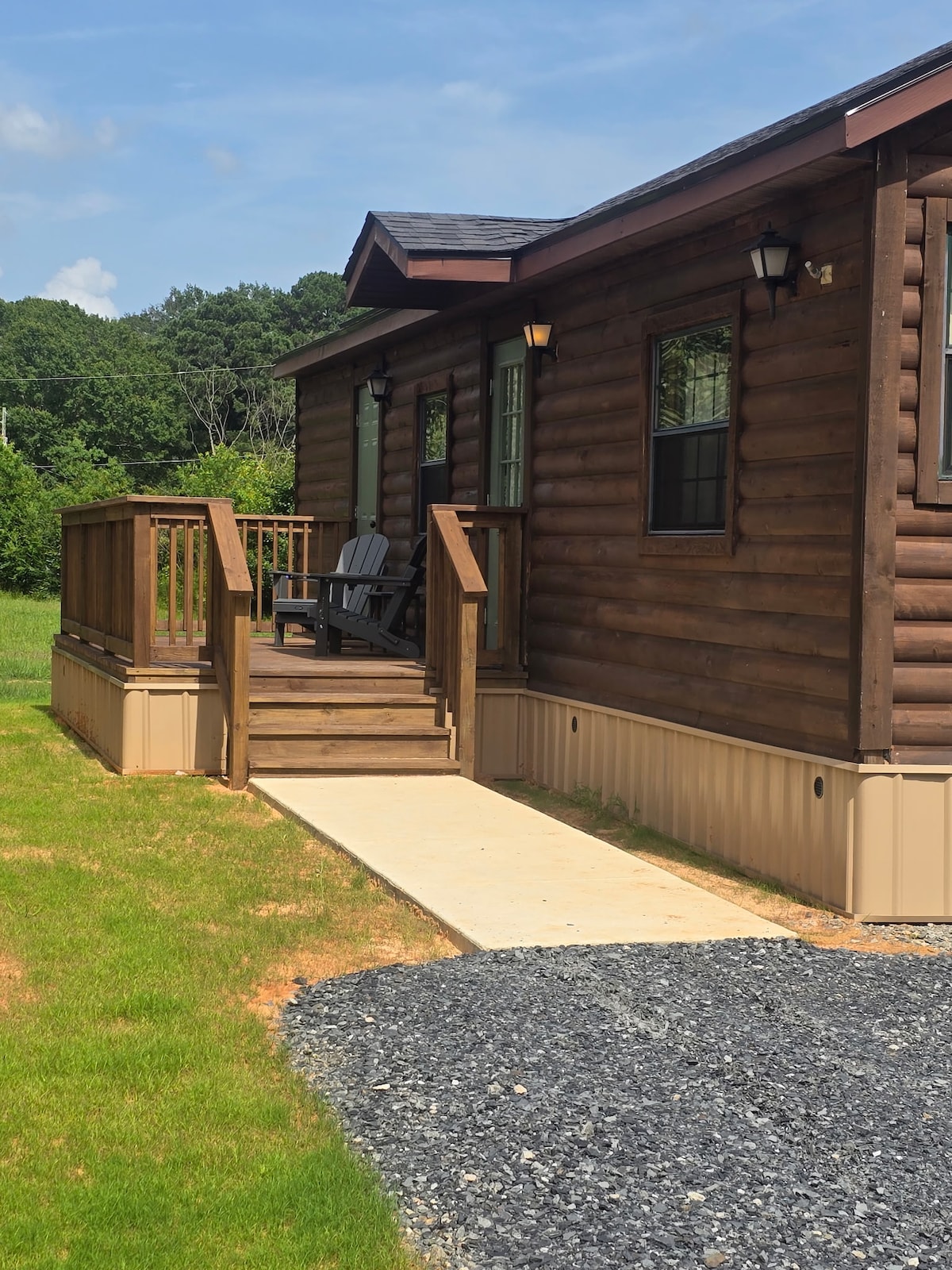 The exterior of a rustic cabin is shown, featuring a wooden deck with Adirondack chairs and sturdy railings. A concrete pathway leads toward the entrance, surrounded by a lawn of green grass, complemented by the natural scenery of trees in the background.