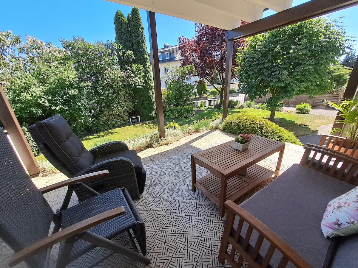 A shaded terrace is presented with comfortable seating arrangements, including a cushioned bench and armchairs. A small wooden table is positioned in the center. Lush greenery and well-maintained plants surround the outdoor space, contributing to a serene environment.