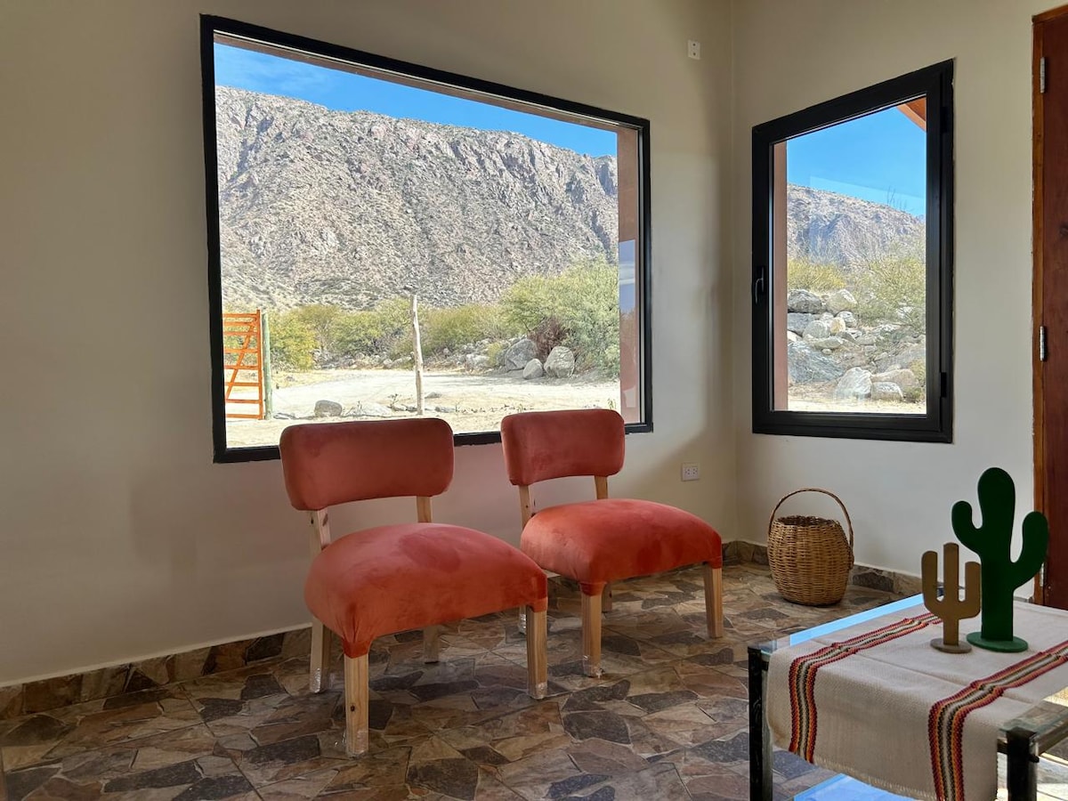 A cozy seating area is presented with two plush chairs positioned near a large window. The view reveals a rocky landscape with natural greenery, on a floor adorned with stone tiles.