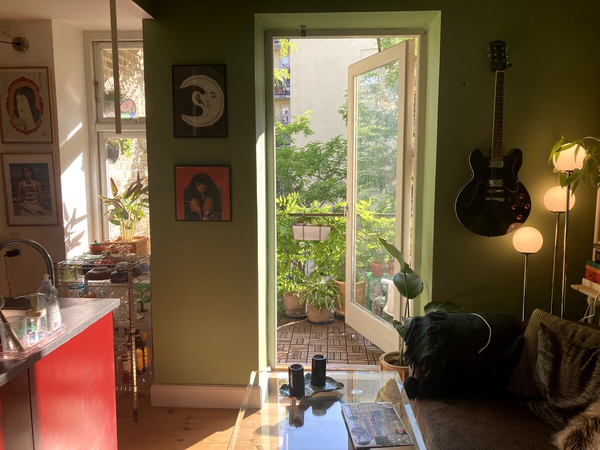 A bright living space features large doors opening to a balcony filled with greenery. A vibrant green wall complements framed artwork. A cozy couch and glass coffee table are visible, alongside an electric guitar mounted on the wall and a plant near the window.