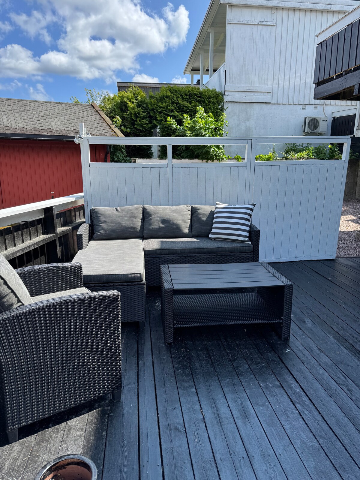 A private outdoor terrace is featured, with a dark wicker seating arrangement consisting of a sectional sofa, two chairs, and a coffee table. A striped cushion adds a touch of contrast. The space is enclosed by a white fence, providing a sense of privacy.
