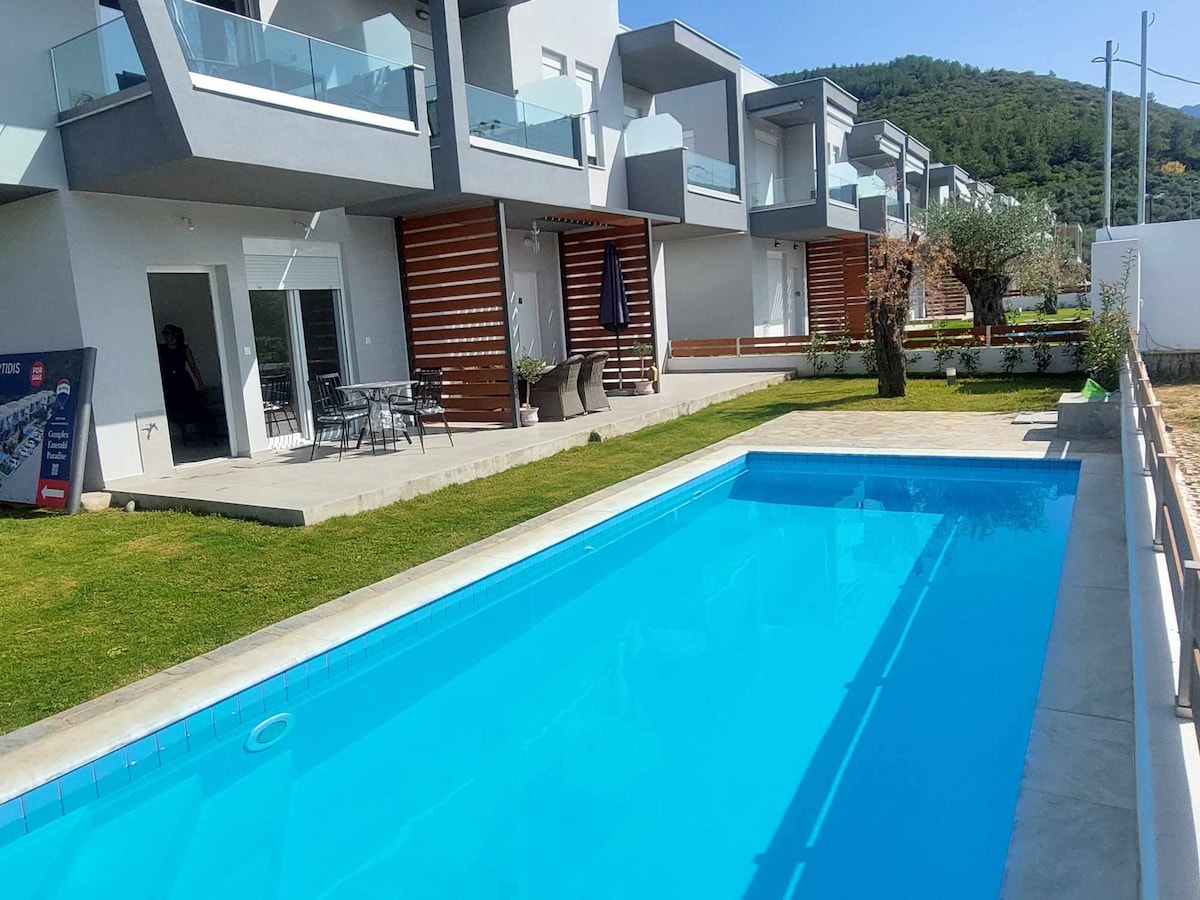 A serene outdoor area features a clear blue pool surrounded by well-maintained grass. Adjacent to the pool, a patio area with tables and chairs is visible, offering shaded seating. The modern building, with multiple balconies, is positioned in the background, framed by lush green hills.