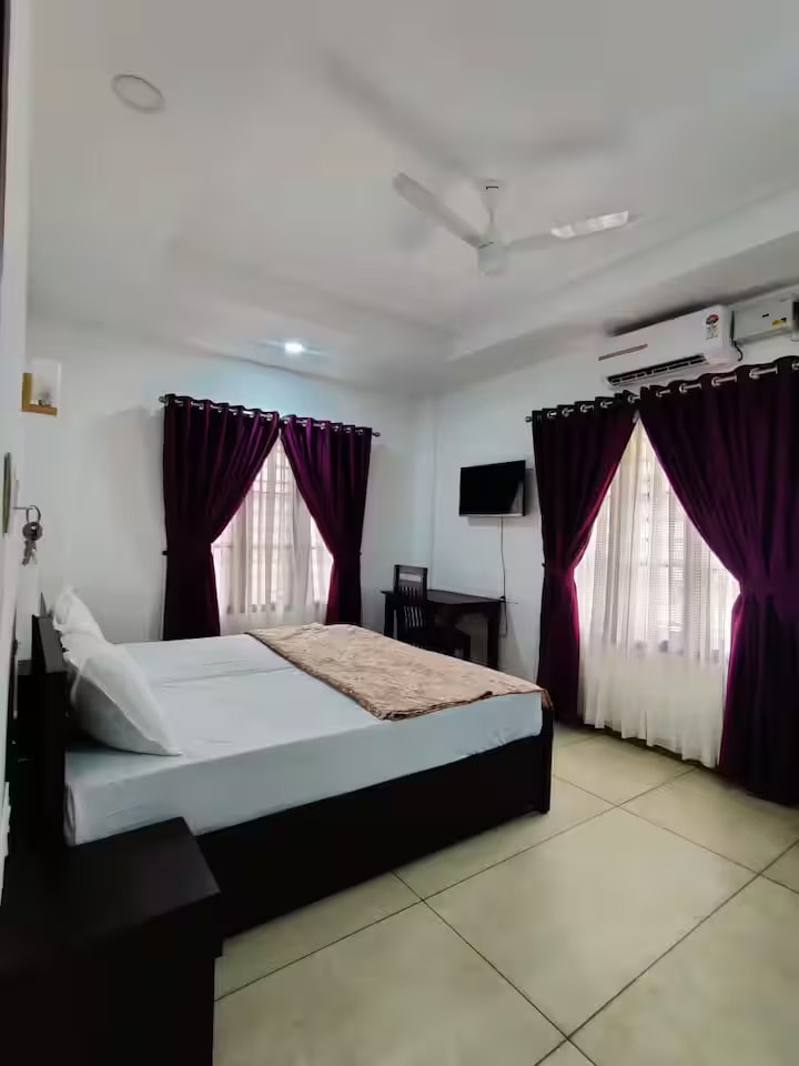 A well-lit bedroom features a spacious bed with a light-colored linen. Dark purple curtains frame a window, allowing natural light to enter. A small desk is situated against the wall, and a wall-mounted television is visible above it. A ceiling fan is installed for comfort.