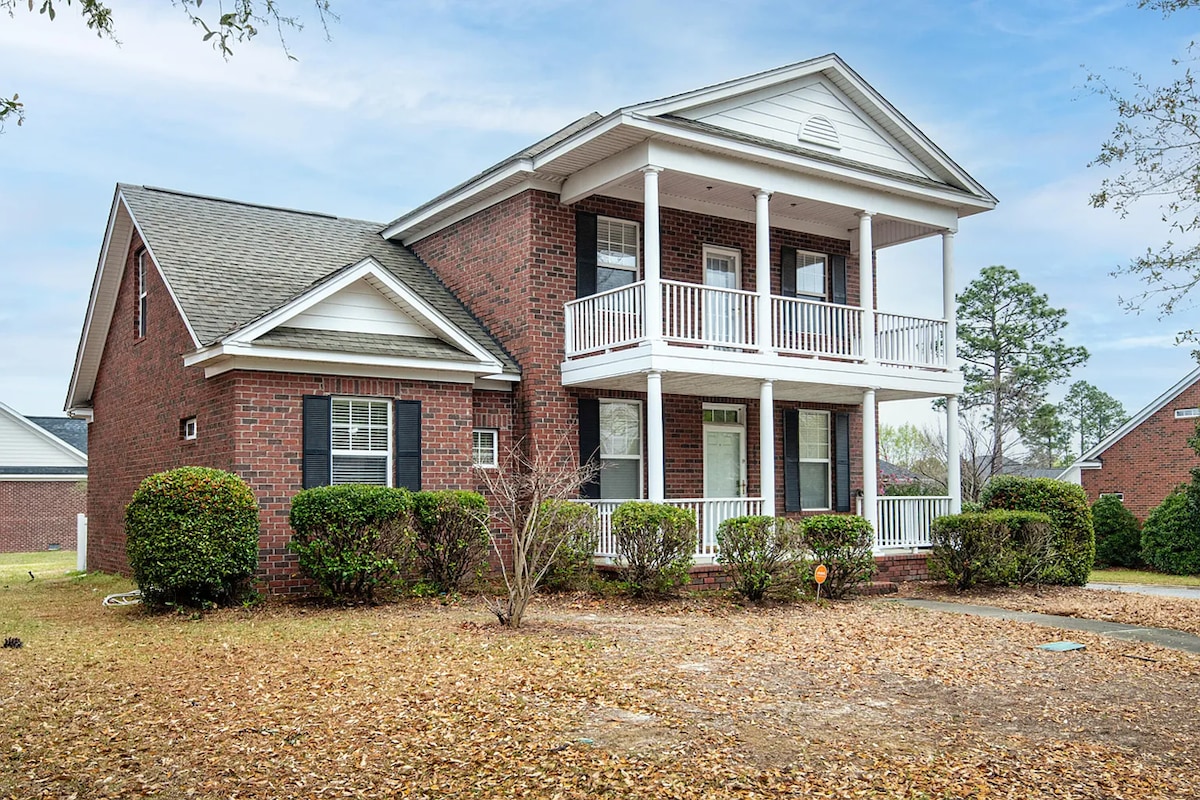 A two-story home features a classic colonial design, with a brick exterior and a welcoming front porch. Lush greenery surrounds the property, and a pathway leads to the entrance, while fallen leaves are scattered across the yard, suggesting a serene outdoor environment.