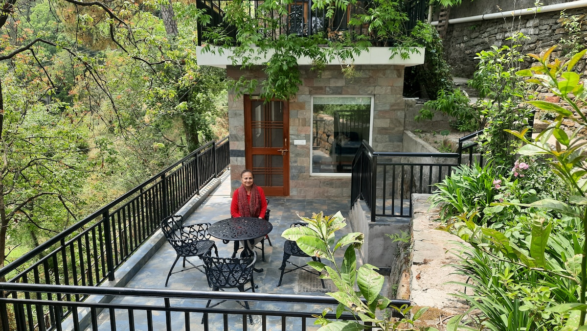 A terraced garden area features a stone and metal table surrounded by four ornate chairs, set against a backdrop of lush greenery. The surrounding plants and trees frame the space, conveying a sense of privacy and natural beauty.