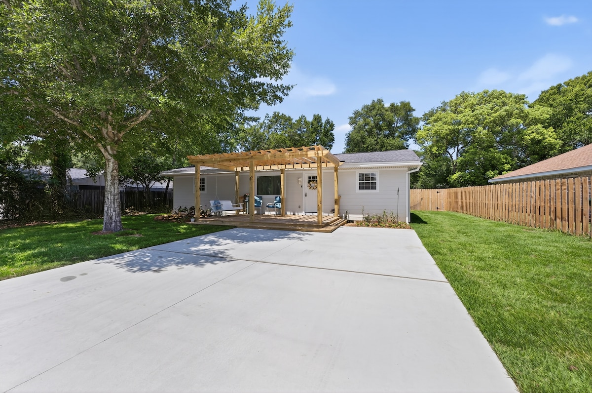 The exterior of a single-story home is showcased, featuring a spacious driveway and a well-maintained yard. A shaded front porch with a wooden pergola provides seating, and the backyard is enclosed by a wooden fence and greenery.