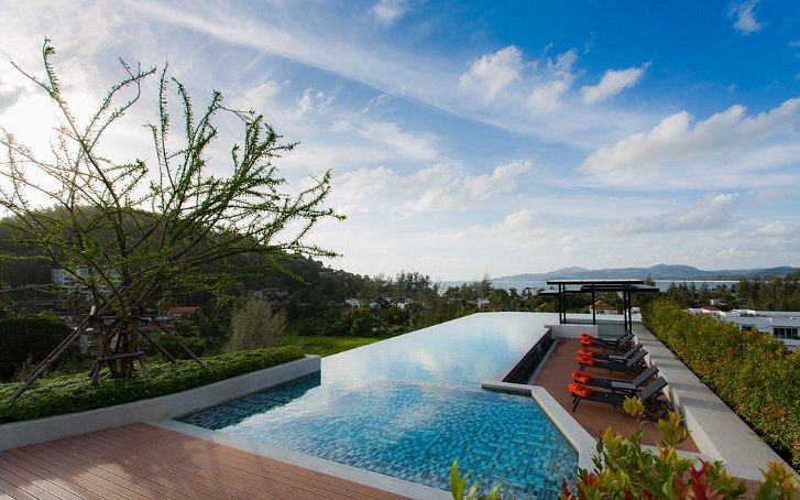 An expansive rooftop swimming pool is visible, framed by lush greenery and scenic hills in the distance. Sun loungers with orange cushions are positioned beside the pool, offering a relaxing spot to take in the surrounding views of the landscape and sky.
