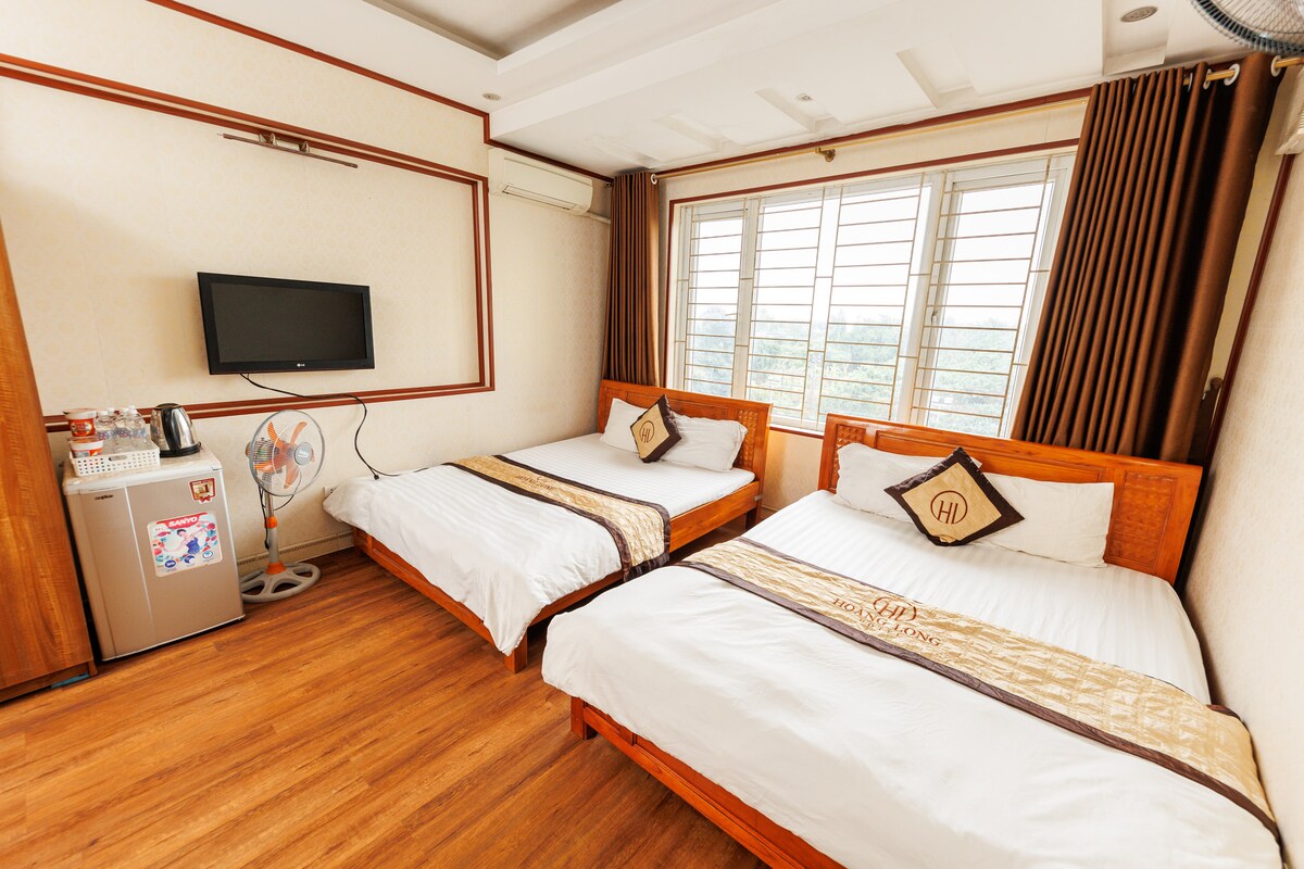 The room features two spacious beds with decorative headboards, complemented by warm wooden flooring. A flat-screen television is mounted on the wall opposite the beds, while a mini-fridge and a kettle are visible in the corner, along with a window providing natural light.