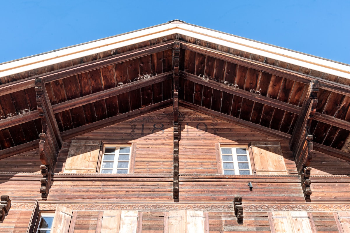 The exterior of a charming chalet from the 1800s is highlighted, showcasing traditional wooden beams and decorative elements under a clear blue sky. Two windows are visible, flanked by wooden shutters that reflect its historical architecture.