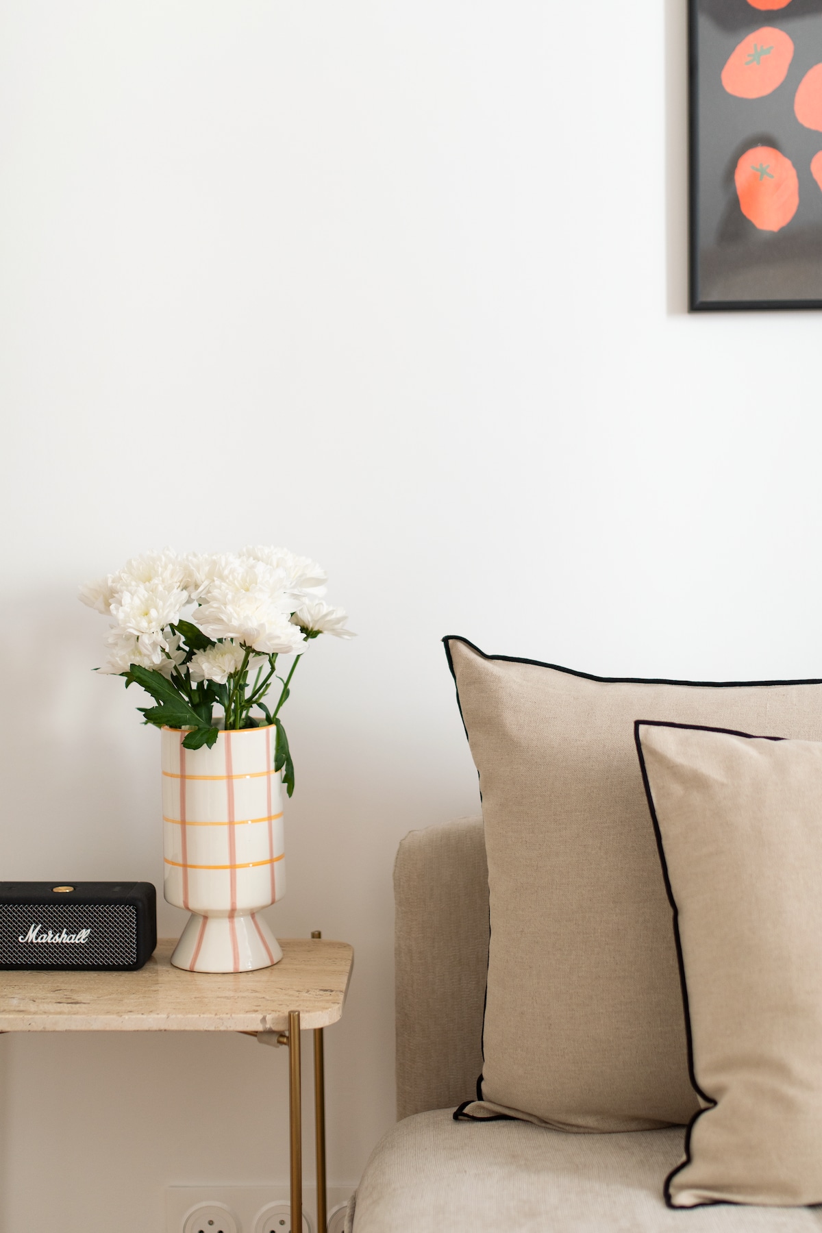 A small side table is positioned next to a comfortable sofa featuring beige pillows with black piping. A vase of white flowers adds a touch of freshness, while a compact Marshall speaker rests nearby, contributing to a stylish and functional living area.