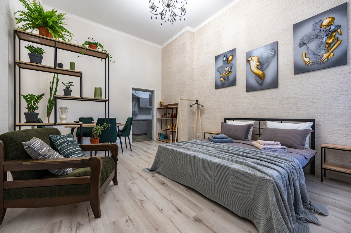 The room features a spacious bed dressed in soft linens, accompanied by an armchair with decorative pillows. A shelving unit with plants adds a touch of greenery, while three framed artworks adorn the walls. A dining table and chairs are visible in the background.