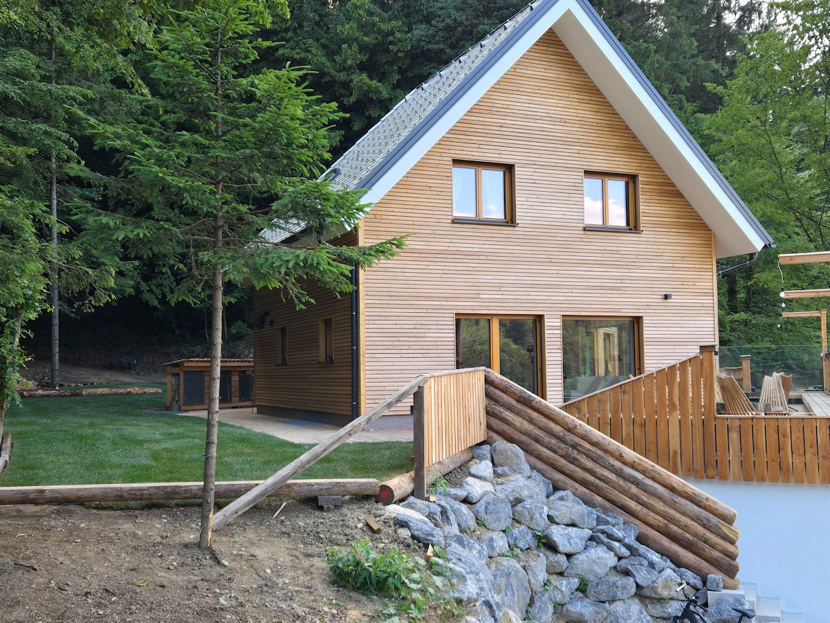 A modern wooden house is nestled among trees in a serene setting. Large windows allow natural light to fill the space, and a wooden deck extends from the building. A gentle slope leads to a stone border, enhancing the natural landscape.